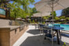 Gas grill area with 2 tables with umbrellas and chairs poolside.